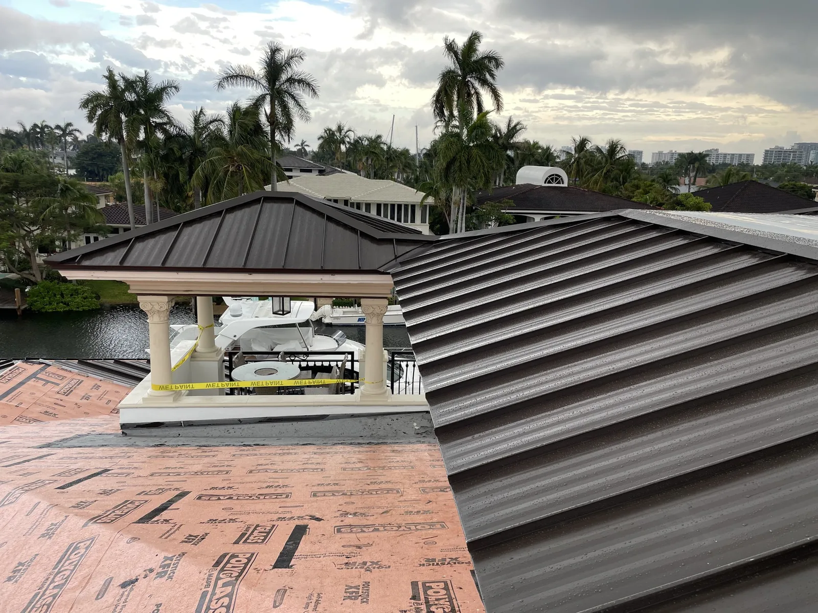 Metal roof panel installation over a waterfront property lined with palms.