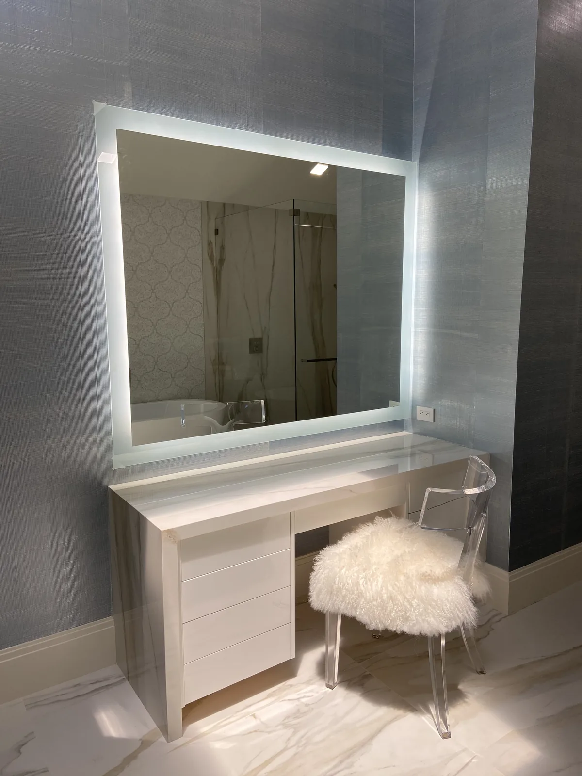 Bathroom vanity with illuminated mirror, white cabinetry, clear acrylic chair.